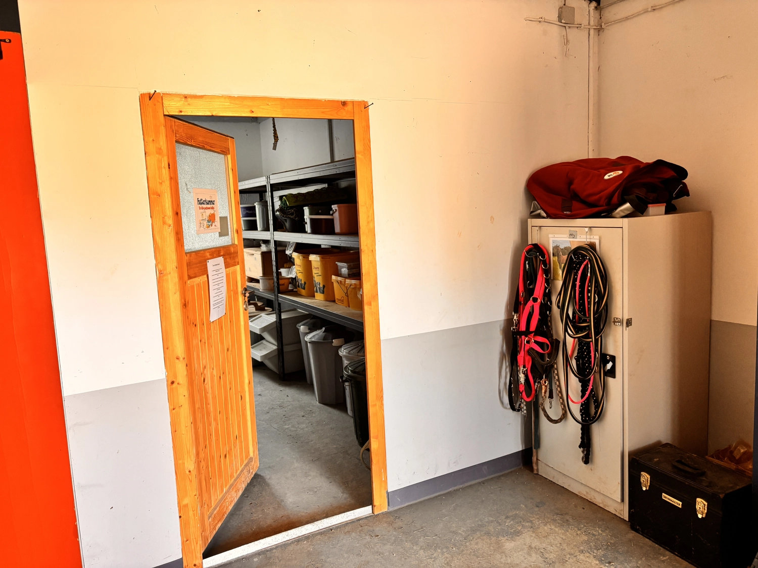Seelenanker Pferde - Pferdetrainer und Reitpädagoge - Eine offene Holztür führt in einen Abstellraum mit Metallregalen. Draußen an der Wand steht ein weißer Schrank, an dem Seile hängen und auf dem eine rote Tasche steht.