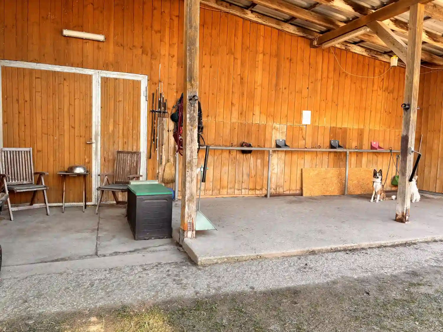 Seelenanker Pferde - Pferdetrainer und Reitpädagoge - Eine hölzerne Terrasse oder ein Stallbereich unter einem Wellblechdach mit einer senkrechten Bohlenwand, Stützpfosten und gestapelten Stühlen.